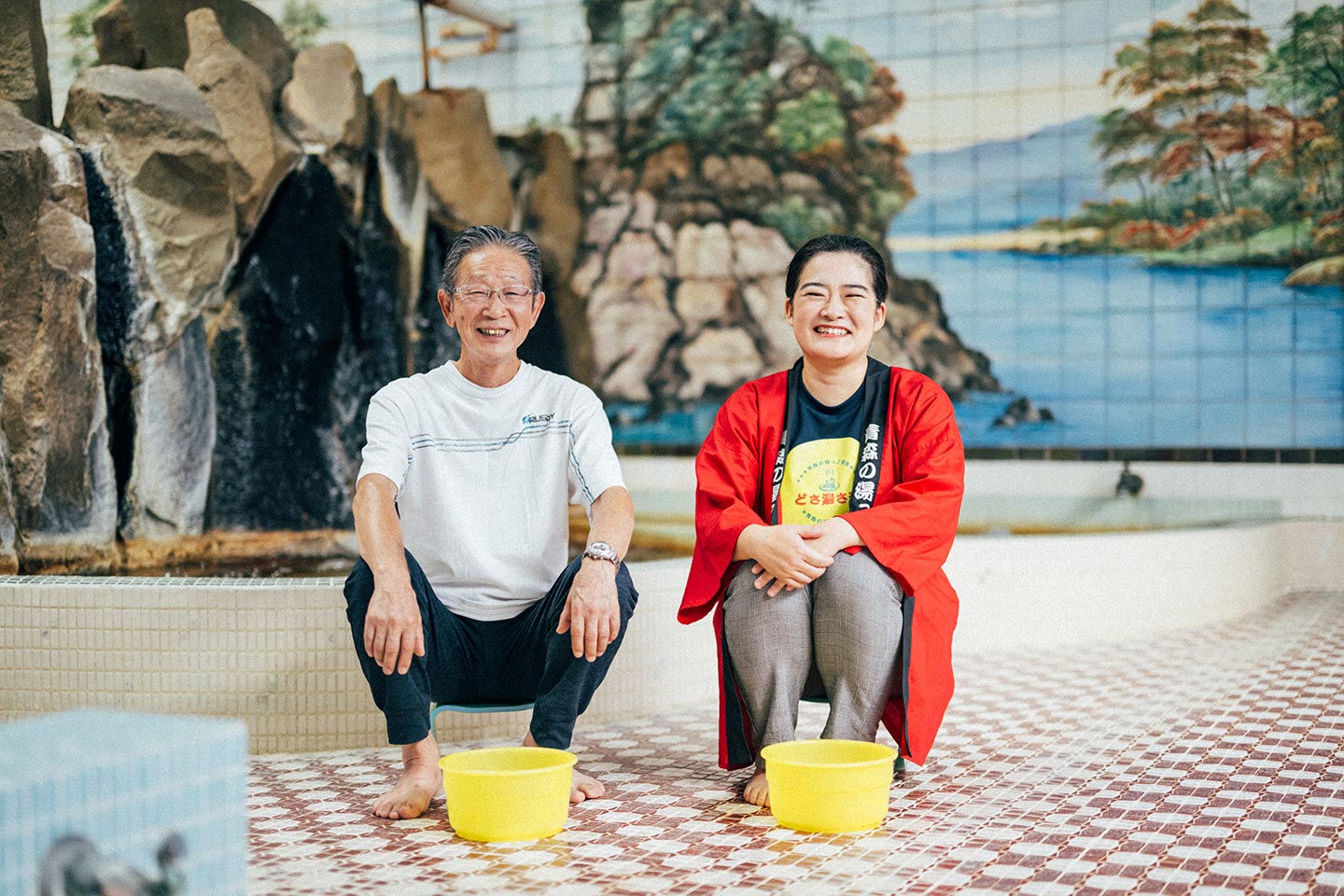油川温泉浴室内で、黄色のお風呂桶の前に並んで座る青森の湯っこ協会・沓掛さんと、油川温泉の佐藤さん