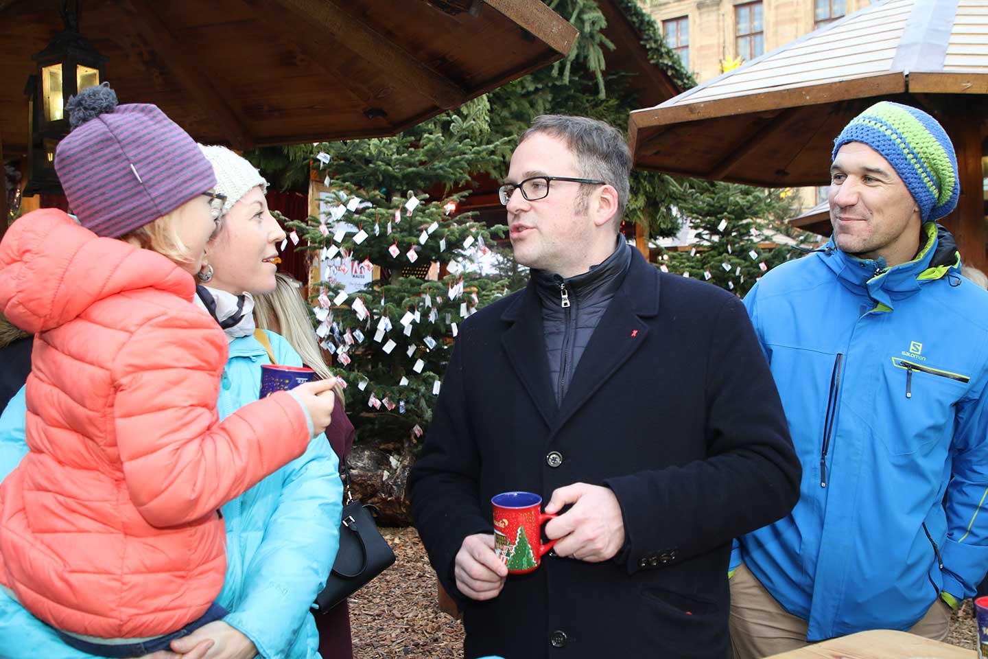 選挙に出る候補者が、クリスマスマーケットの一角で飲み物を片手に市民と対話している様子。男性と子どもを連れた女性が話している