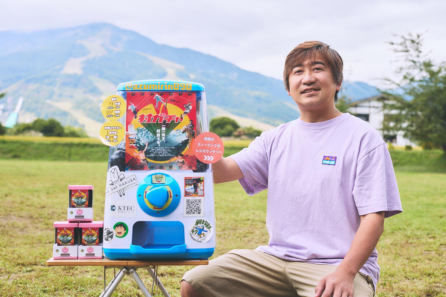 芝生の広場で、村ガチャの筐体の横に座る佐藤敦俊さん