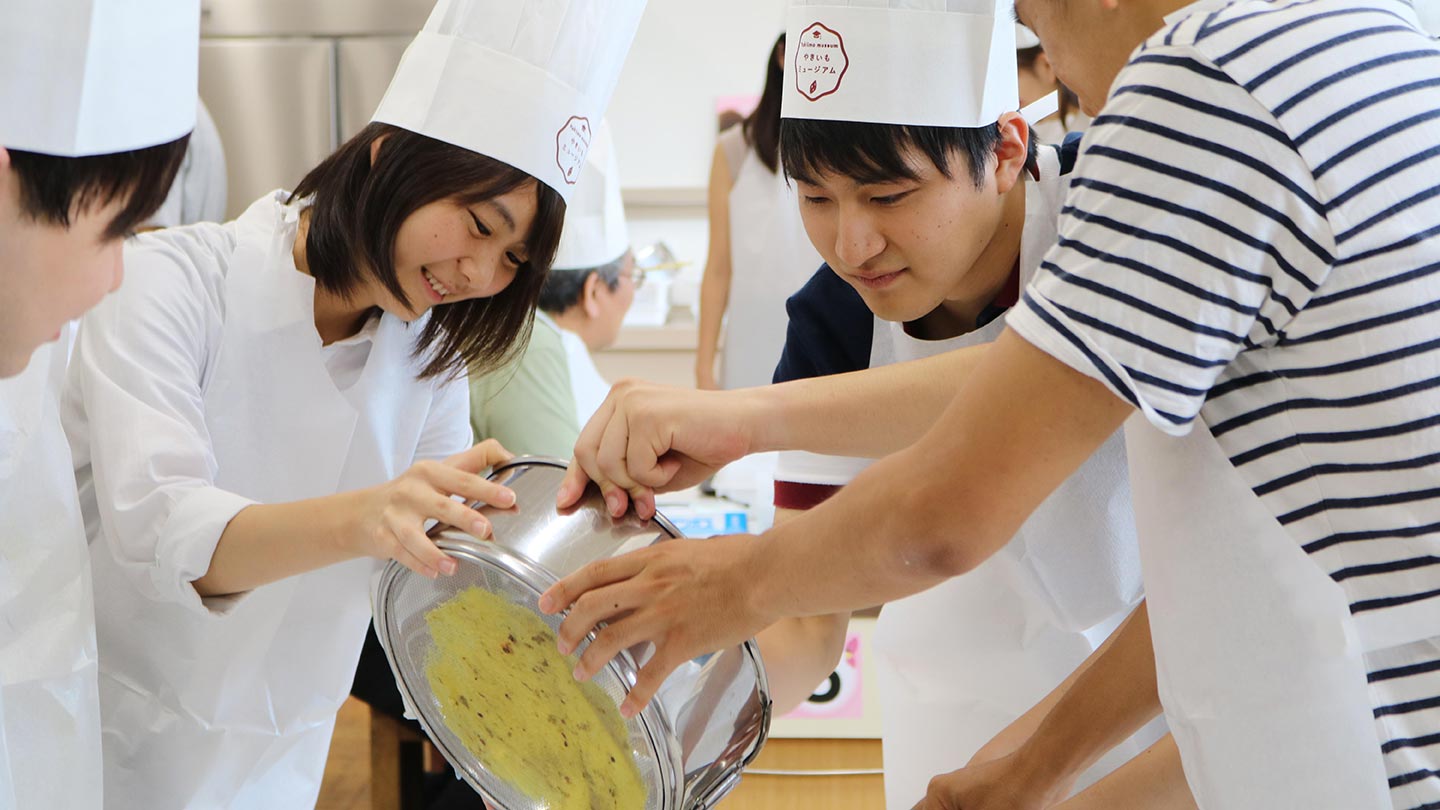 エプロンとコック帽子を身につけた子どもたちが、3人がかりでサツマイモを裏ごししてお菓子をつくっている