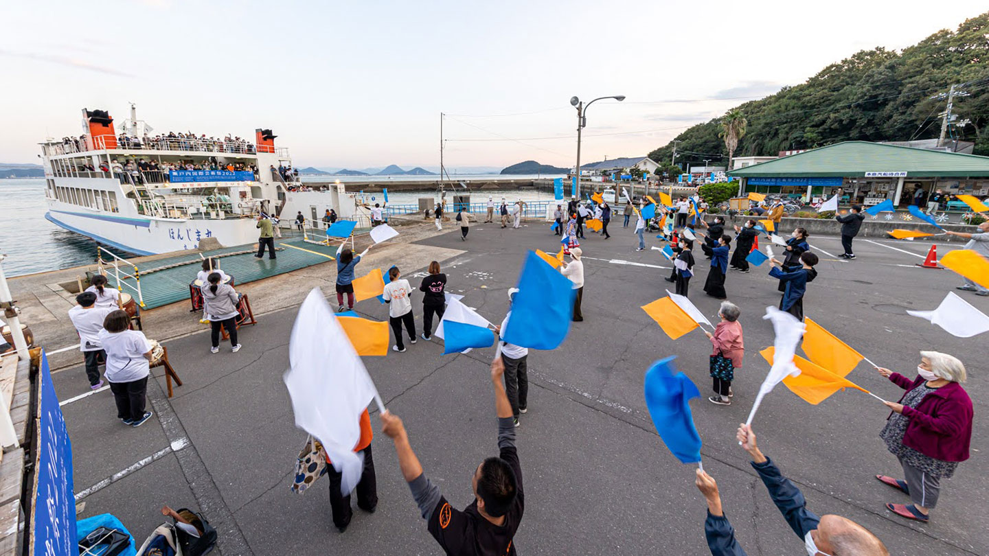 港で船を見送る人々。老脈男女が白や青、オレンジのカラフルな旗を振っている