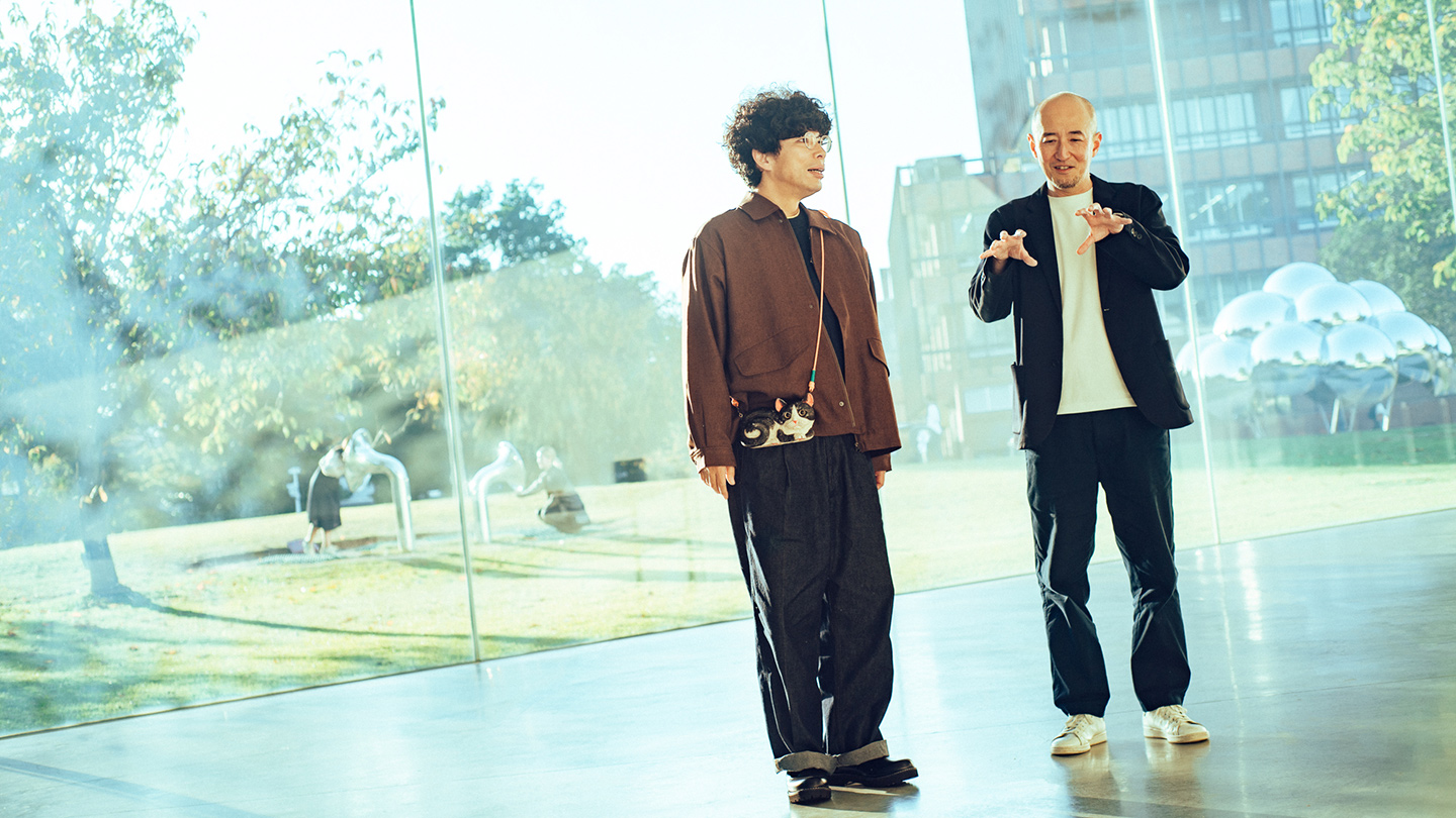 ガラス張りの館内廊下で話す片桐仁さんと鷲田めるろさん。ガラス窓の外側では、芝生の広場で市民がアート作品に触れている