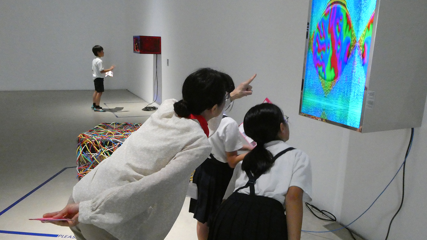 カラフルな映像が画面に映し出されているデジタルアート作品の前で、大人がその作品に指をさしながら、生徒2名に解説している様子