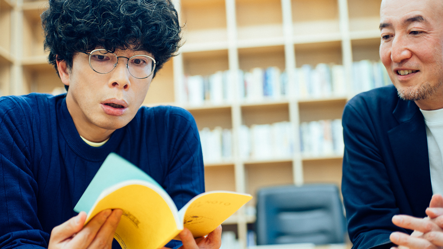 『ROAD TO NOTO NOTE』のページをめくる片桐仁さんと、そのノートについて説明する鷲田さん