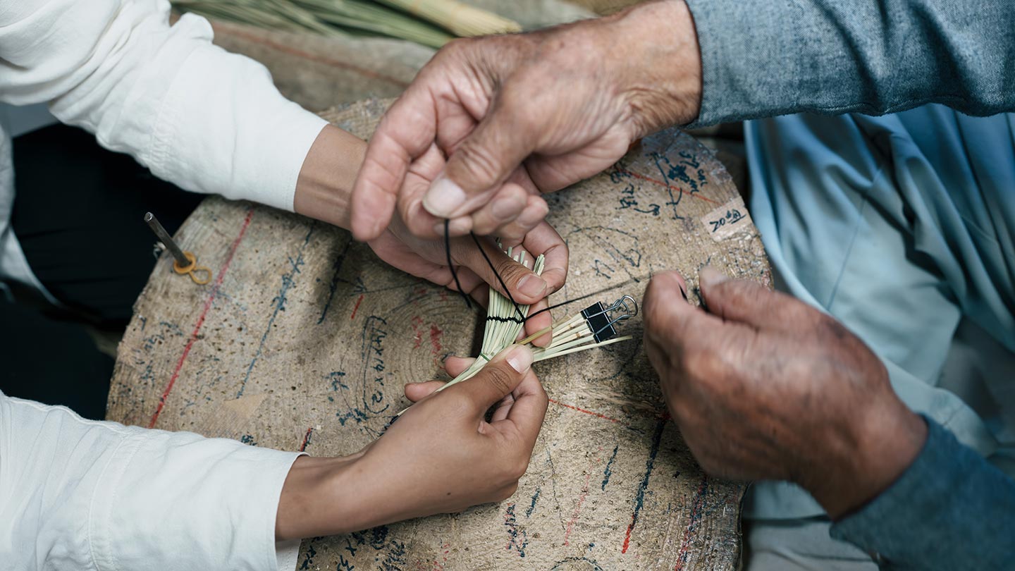 鶴の飾り物を作る工程。わらを押さえる智香さんの手と、わらに黒い糸をかけていく清さんの手