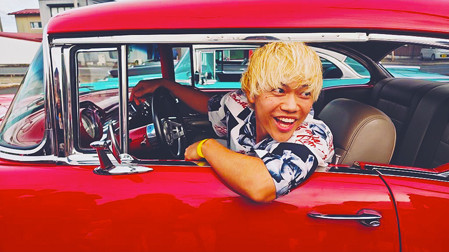 寺中友将さんのポートレート。赤い車の運転席に乗り、左肘を窓にかけ、車の外に視線を向けている