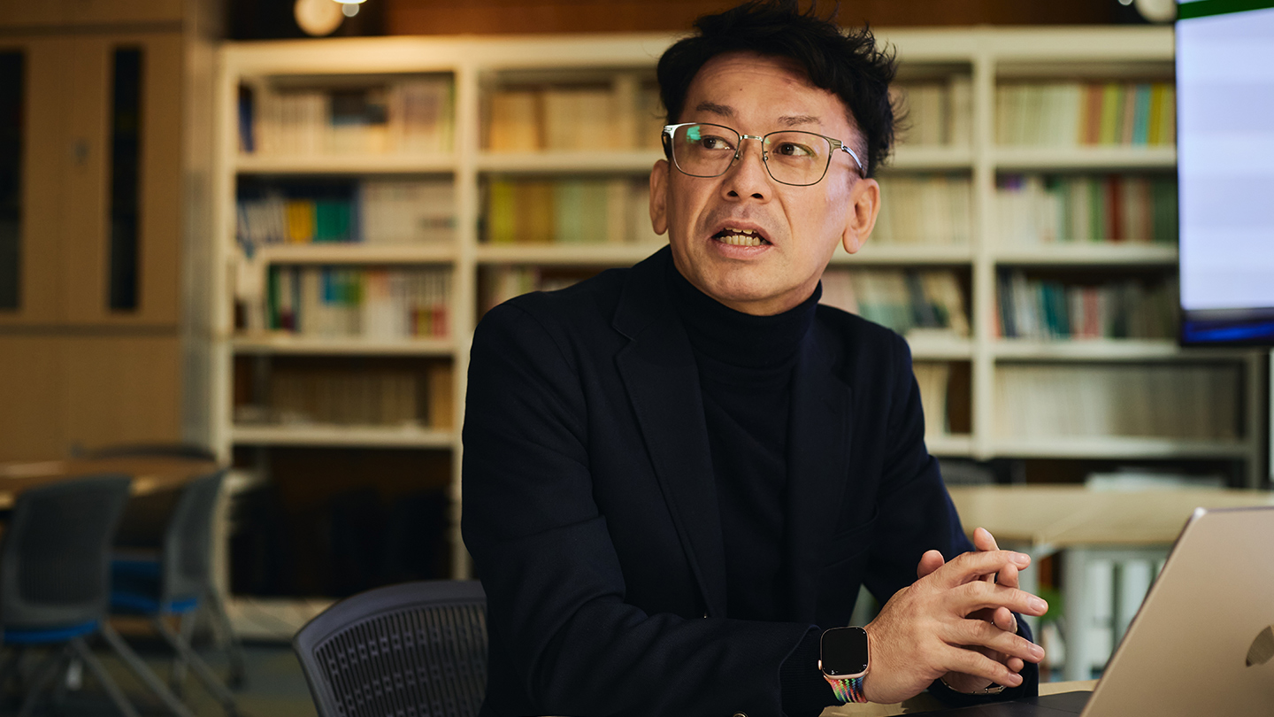 書物がたくさん並んだ本棚を背景に、話している佐藤さん