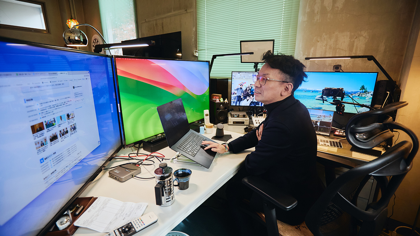 佐藤先生が研究室の椅子に座って、デスクの上に5台並べられた大小の画面を見ている