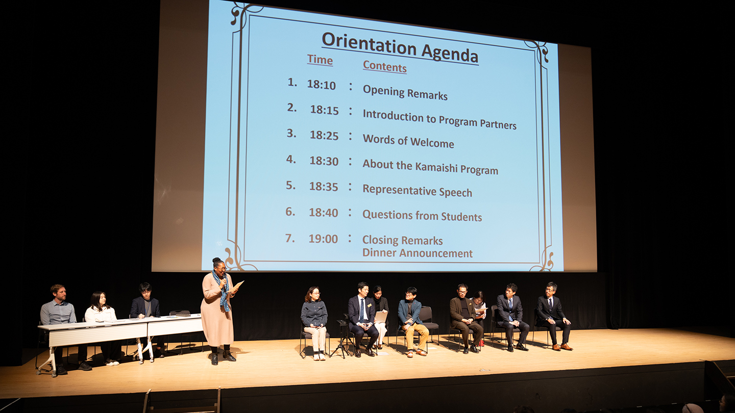 大きなスクリーンにアジェンダが映し出され、壇上に人が何名か椅子に座っている