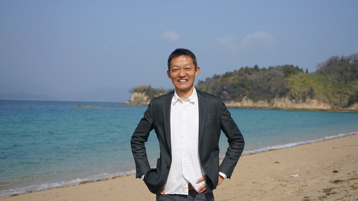 取釜宏行さんのポートレート。海を背景に砂浜で微笑んでいる