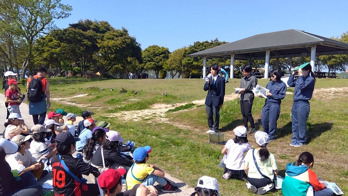 海草をふやすキットを小学校に展開した生徒が4人で、子どもたちに新しいプロジェクトの説明をしている。屋外教室で、広場で行われている