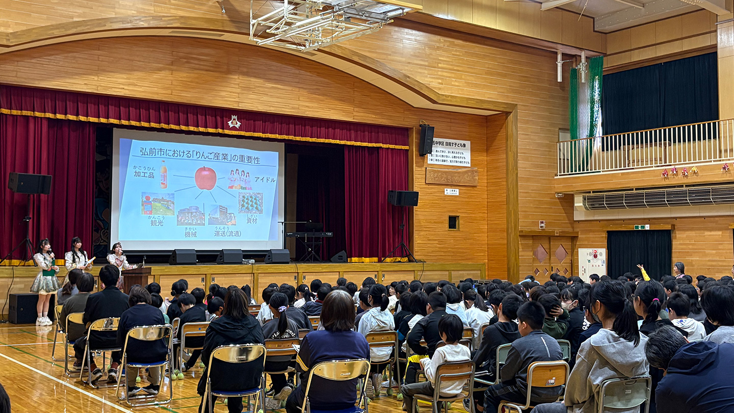 小学校の体育館で、子どもたちの前で授業をするりんご娘