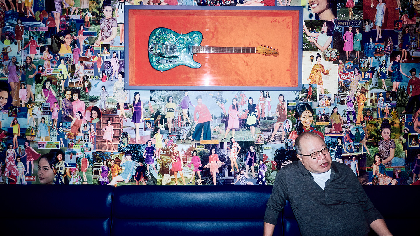 ギターと雑誌の切り抜きが飾られた壁の前で話す都築響一さん