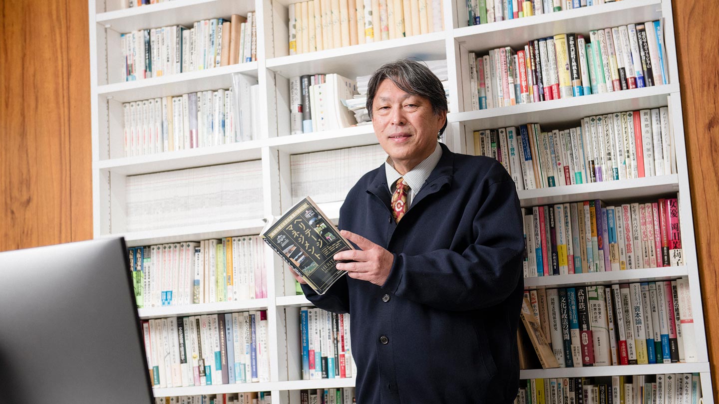 本棚を背にして本を手に持ち立っている文化政策学者の松本茂章さん