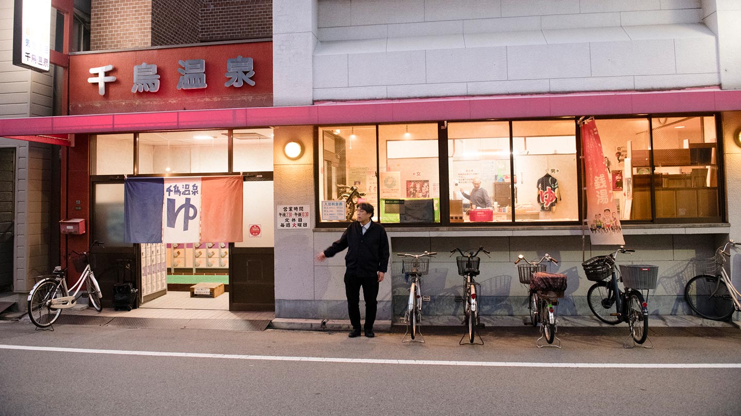 千鳥温泉の外観と、その前に立つ松本さん。自転車が数台置かれている。紺、白、オレンジで構成されたのれんがかけられており、「ゆ」の文字が書かれている