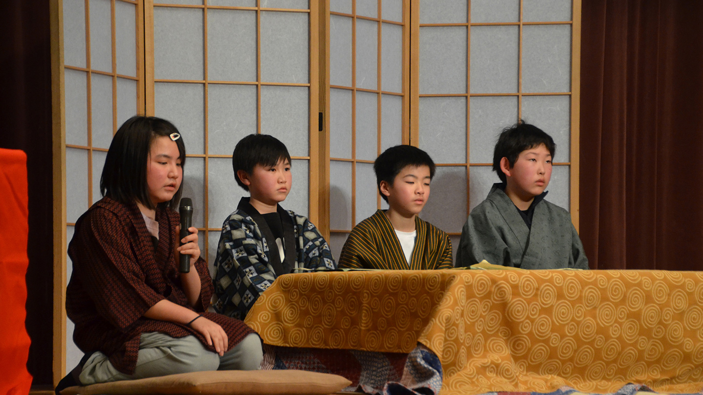 4人の小学生がローテーブルの前に座り、一番左の女の子がマイクを持ち、語りを披露している