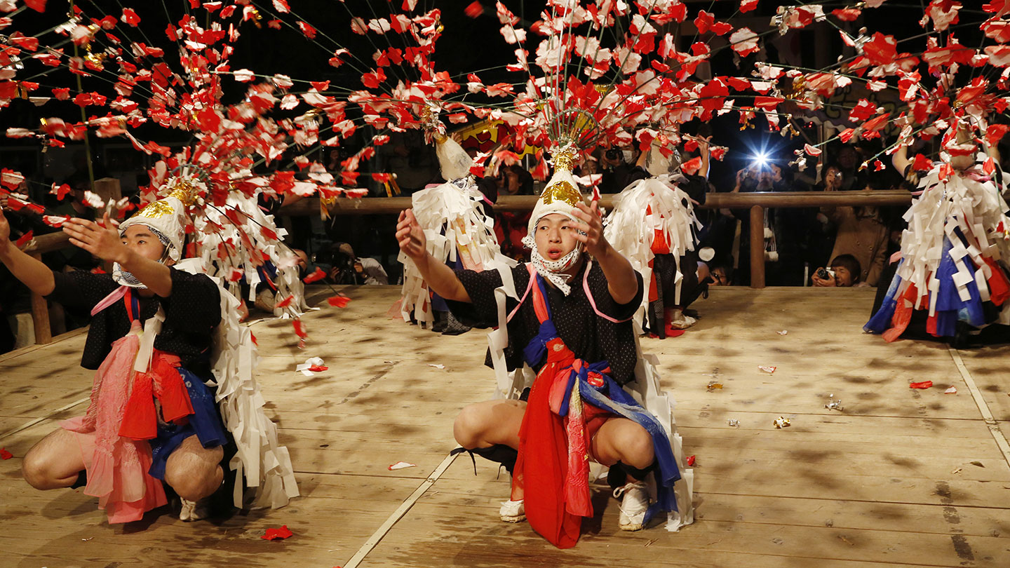 たすきや帯で飾った色彩豊かな衣装を身に着け、華やかな装飾が施された藁笠を被った青年たちが夜の舞台で舞っている