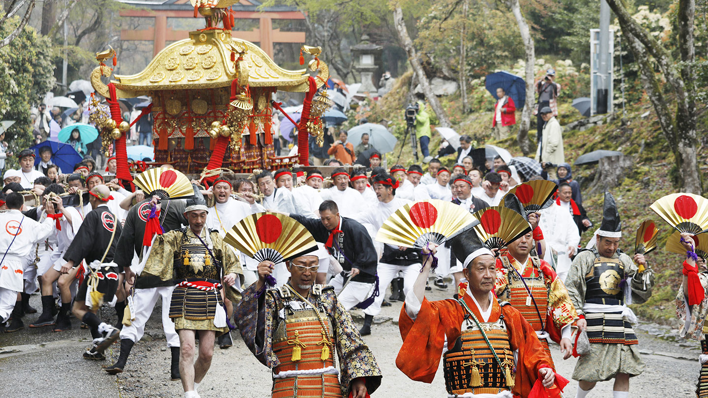 日の丸の扇を掲げ、鮮やかな鎧を着て行進する男性たち。背後には金色の屋根の神輿と赤い鳥居が見える
