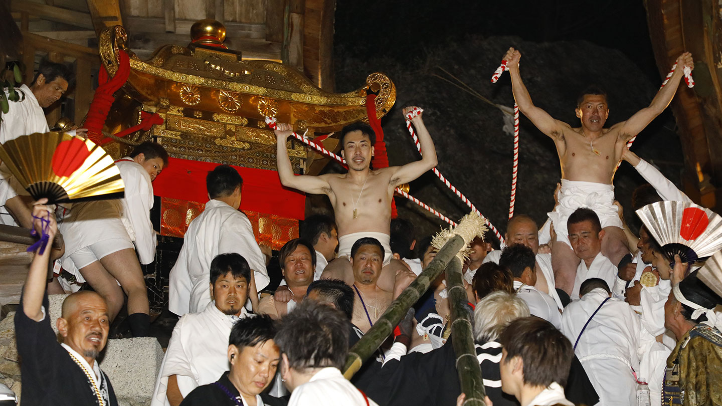 夜の暗闇のなか、神輿を運ぶ人々。肩車された人や、扇を掲げた人もいる