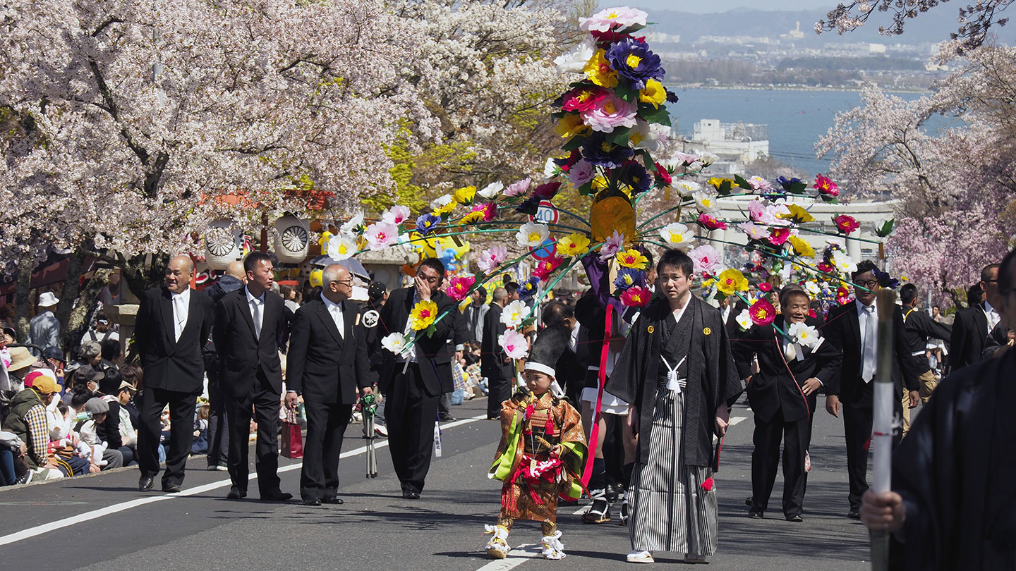 鎧を着た5歳前後の稚児が先頭を歩き、神さまに捧げる花を運んでいる。背後には親類の大人が見守っている
