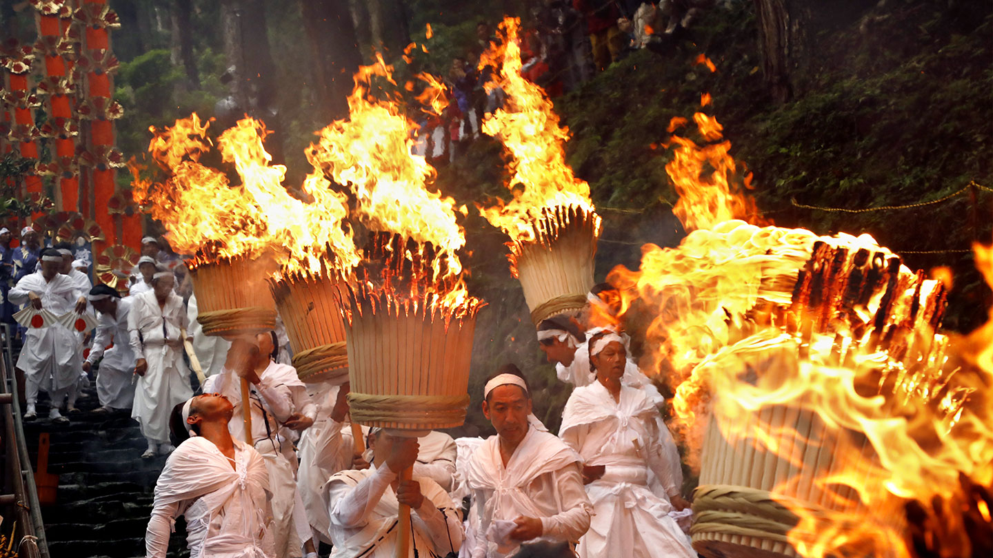 火のついた巨大な松明を運び、白い装束姿で参道を進む氏子たち。奥には赤い扇を重ねた12基の扇神輿が見える