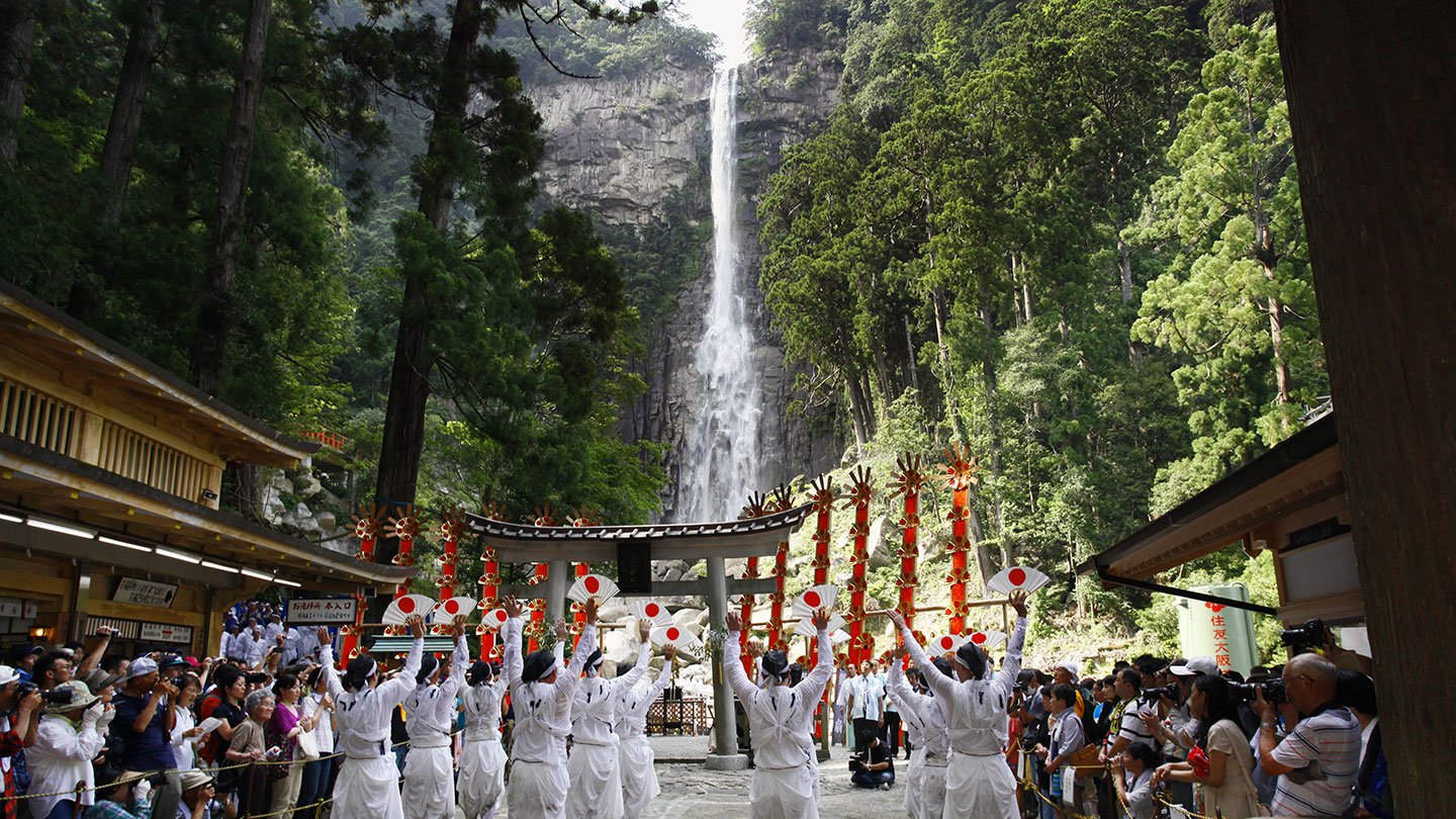 奥には木々に囲まれた大きな滝、手前には扇神輿とそれらに祈る白装束の氏子たち。両脇には観光客の姿