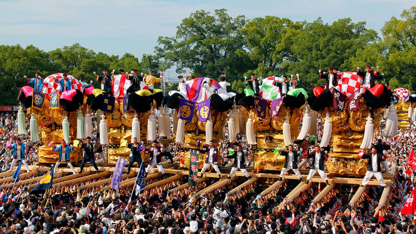晴れた空の下、カラフルな天幕がかけられた巨大な5つの太鼓台と、それらを担ぐ人々