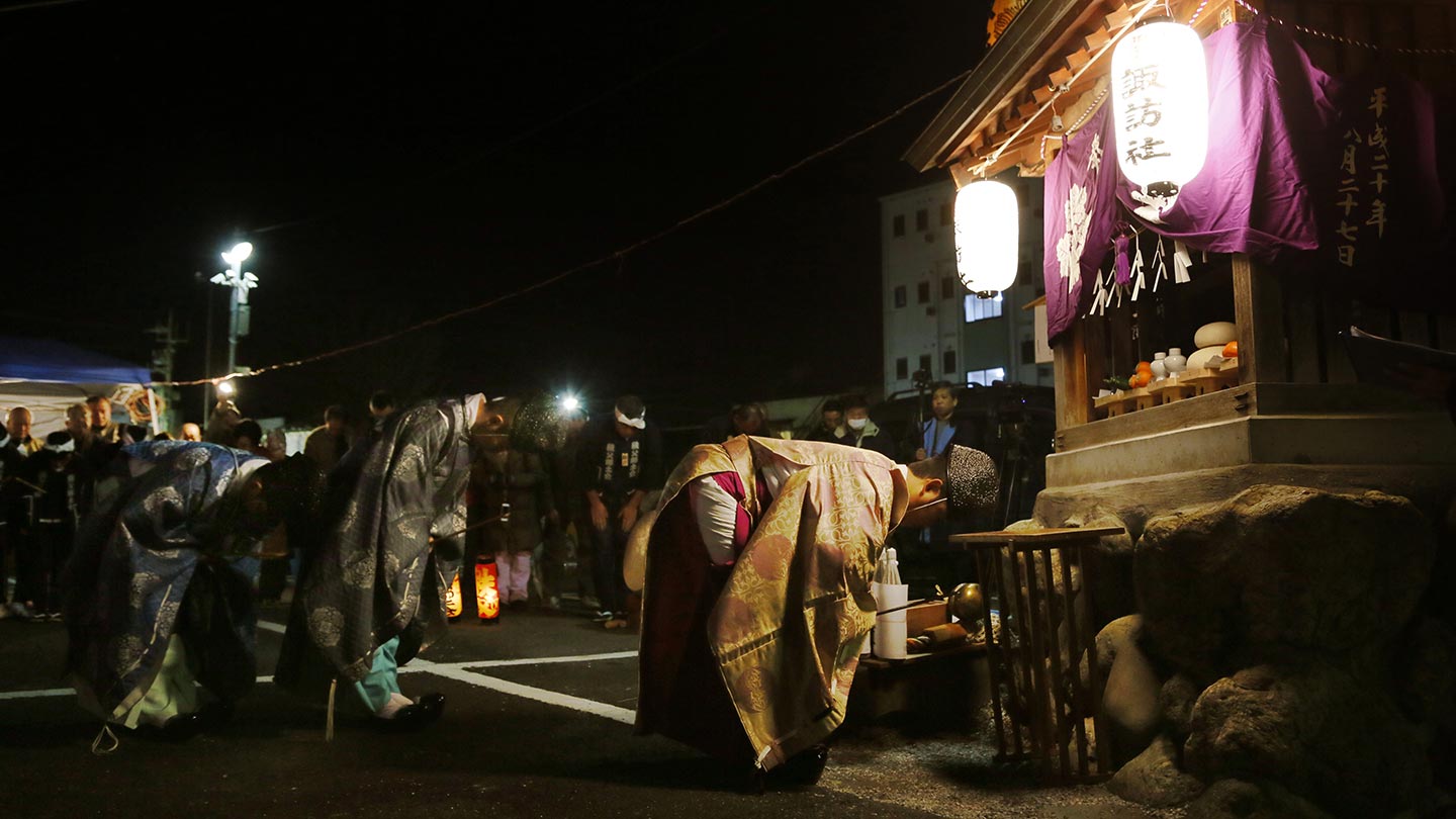 「諏訪神社」と書かれた提灯が掲げられた祠に頭を下げている神職と町の役員