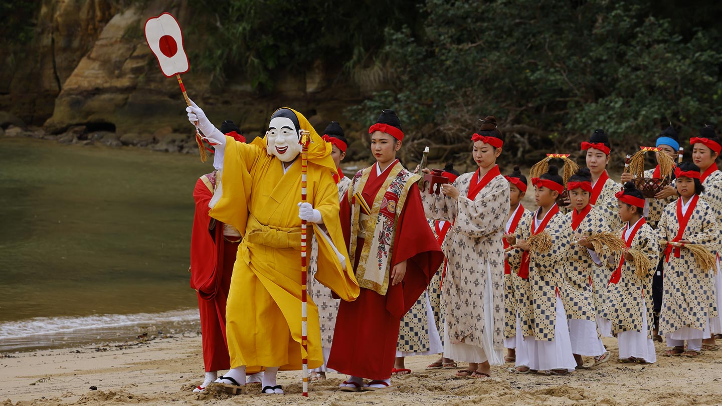 仮面と黄色い衣（ころも）を身につけた「ミリクさま」を先頭に、八重山の伝統衣装を着た人々が浜辺を歩いている