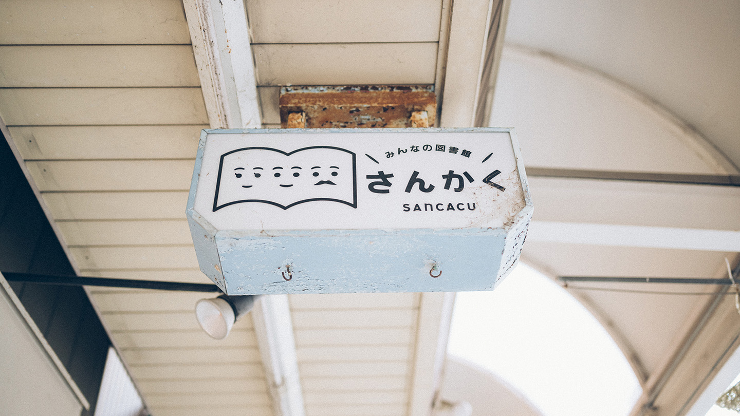 商店街の軒下に取り付けられた「みんなの図書館 さんかく」のロゴ入り看板を見上げた様子