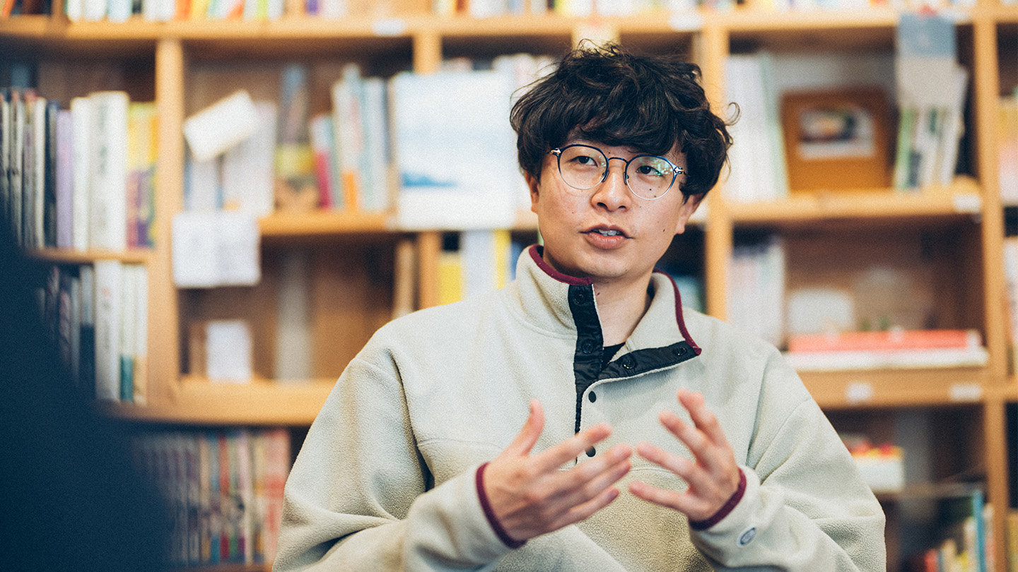 本棚が並ぶ図書スペースで、眼鏡をかけた土肥さんが椅子に座り、手振りを交えて話している様子