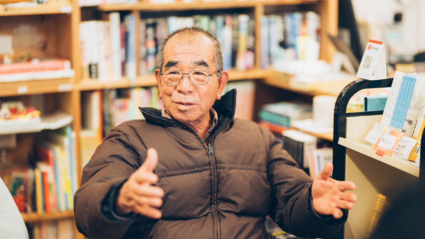 本棚を背景に、椅子に座った栗下さんが身振りを交えて話している様子