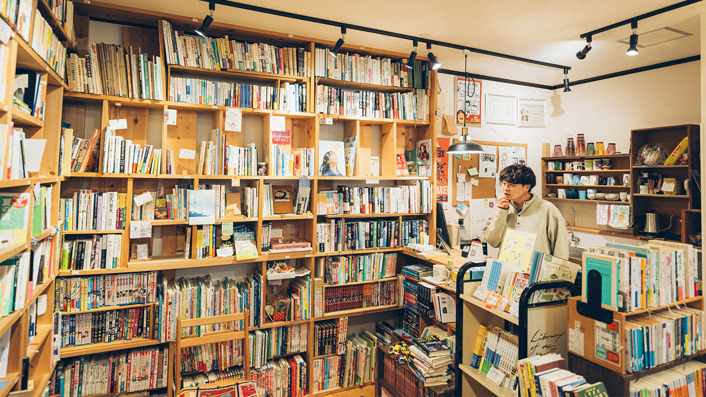 壁一面の木製本棚に本がぎっしり並ぶ図書スペースで、土肥さんがカウンター越しに本を眺めている様子