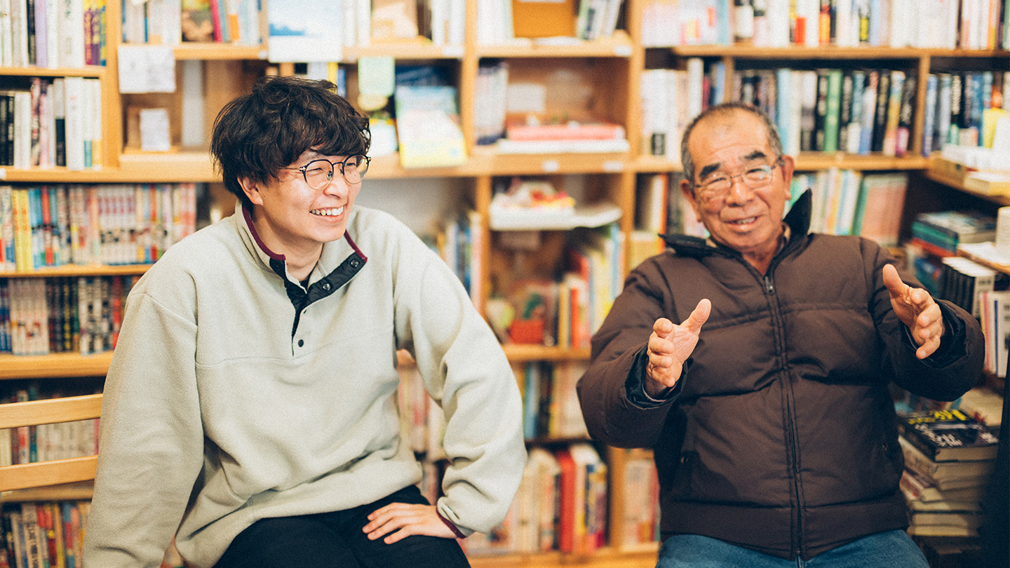 本棚を背景に、土肥さんと栗下さんが並んで笑顔で会話している様子