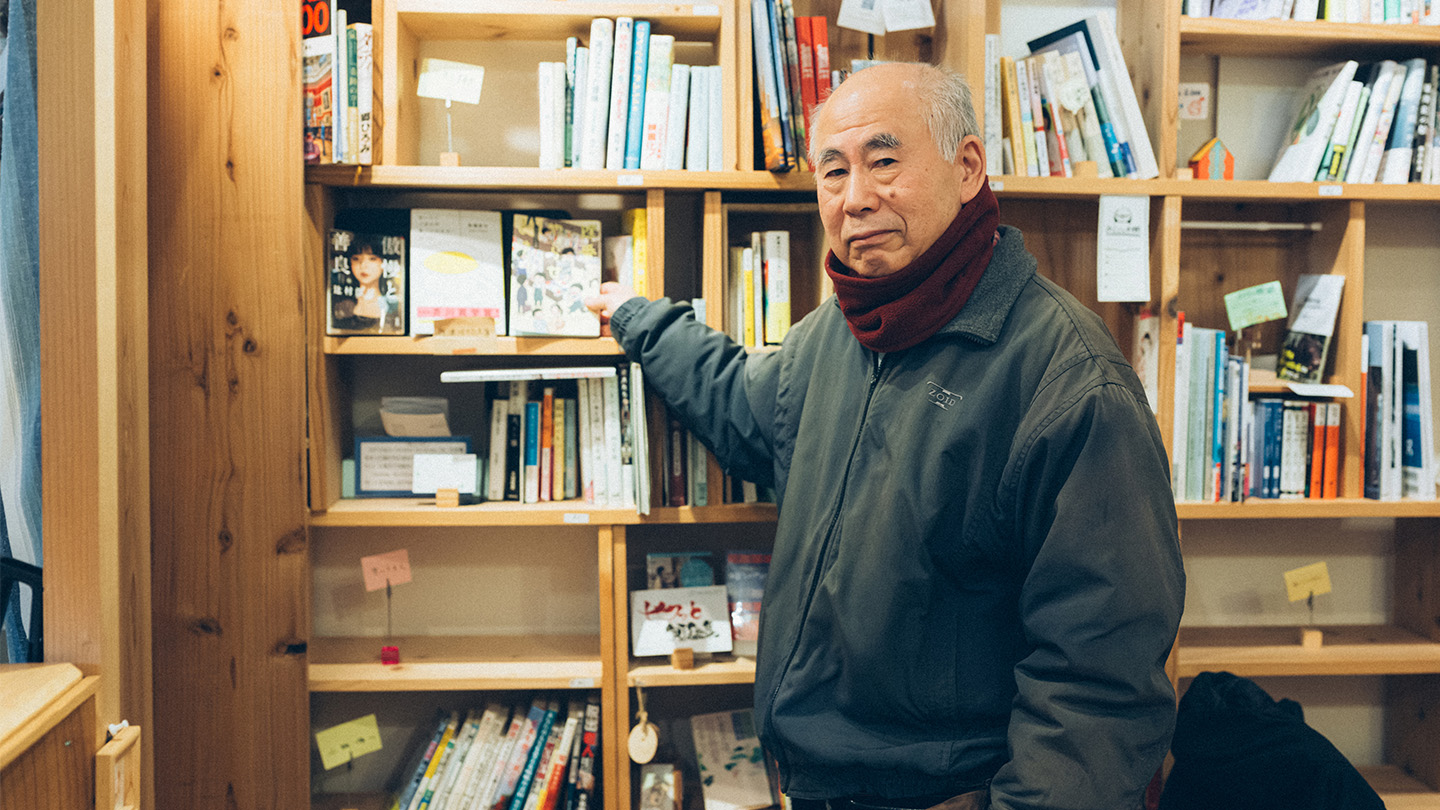 本棚が並ぶ図書スペースで、棚の一角を指さして立っている村松さん