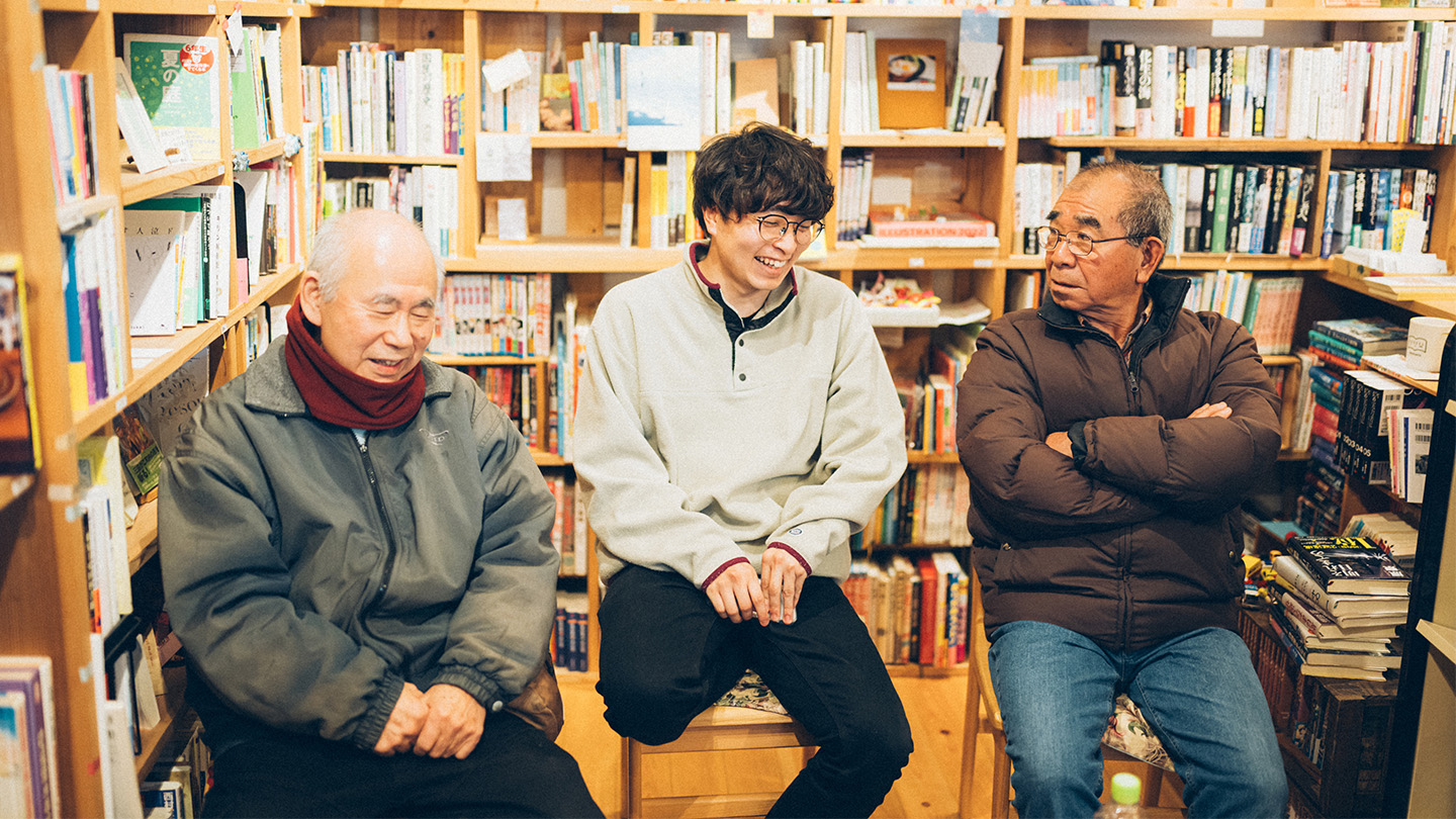 書棚に囲まれた小さな図書館の室内に、村松さんと土肥さん、栗下さんが並んで座り、笑顔で会話している様子