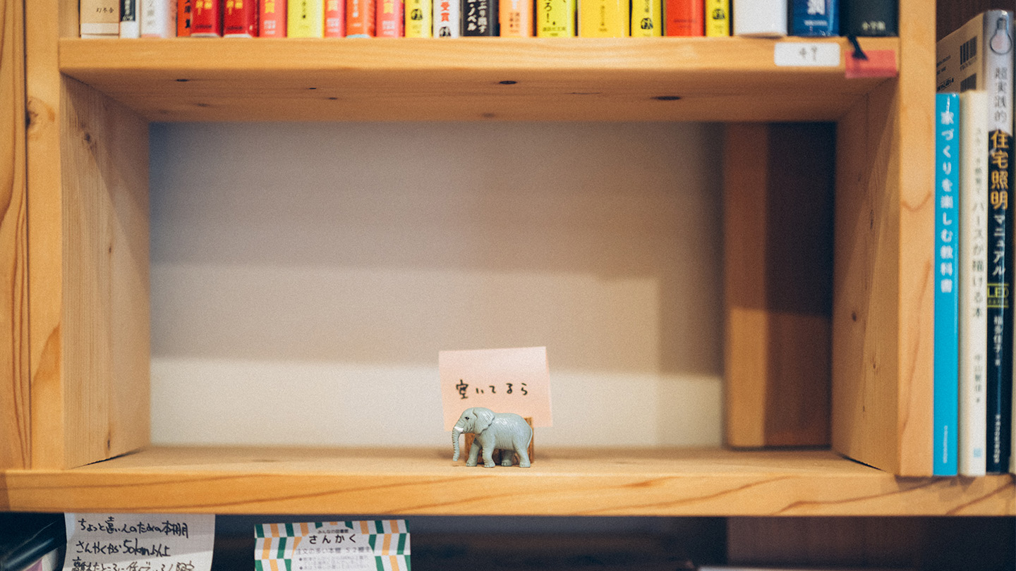 木製の本棚の空いたスペースに、小さなゾウの置物と「空いてるら（＝空いてるよ）」と書かれたメモが置かれている様子。周囲には本が並び、棚の一部が空席であることを示している