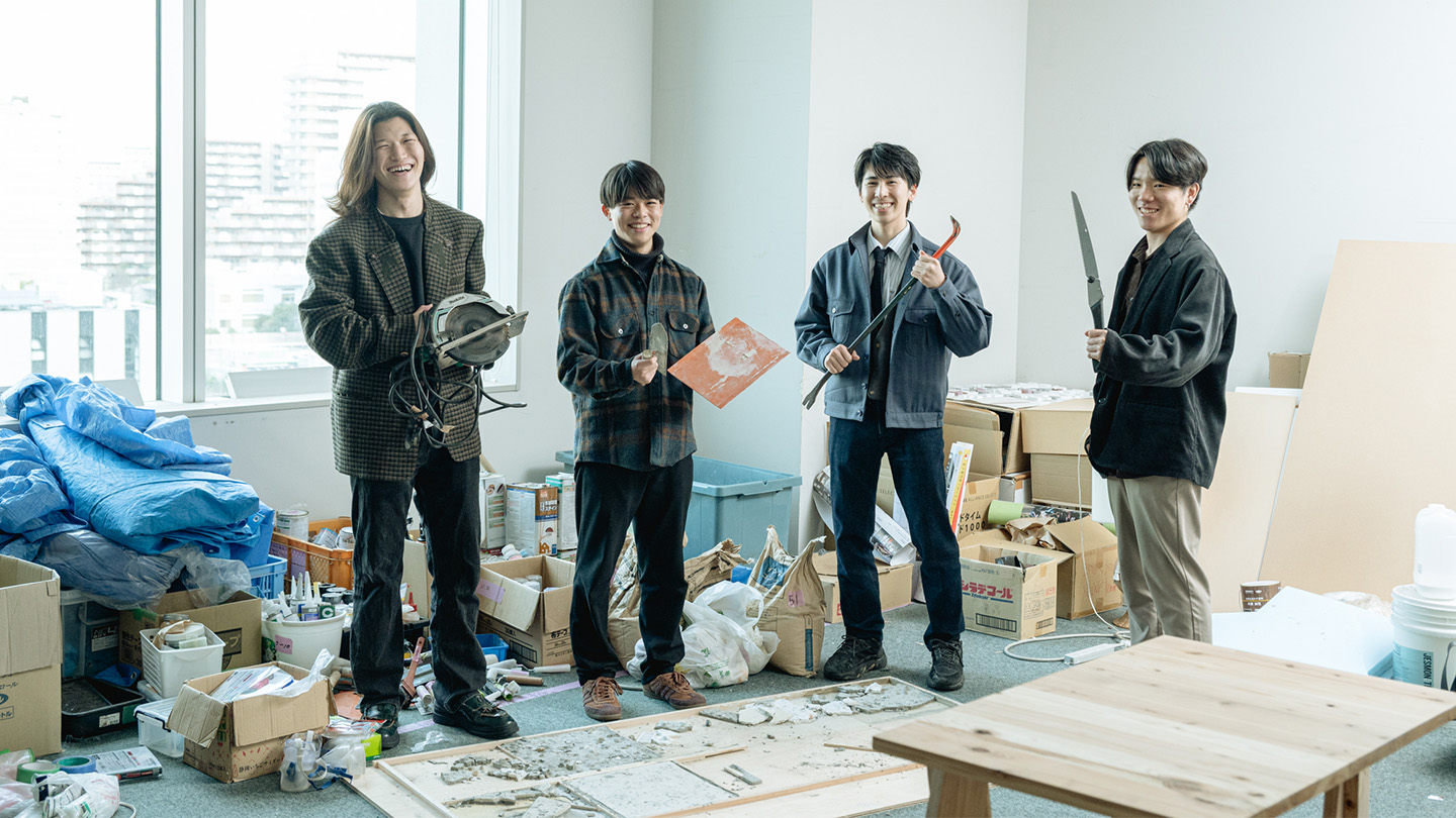 模型や作業台、工具や資材で雑然とした部室のなかに立つ男子学生4名