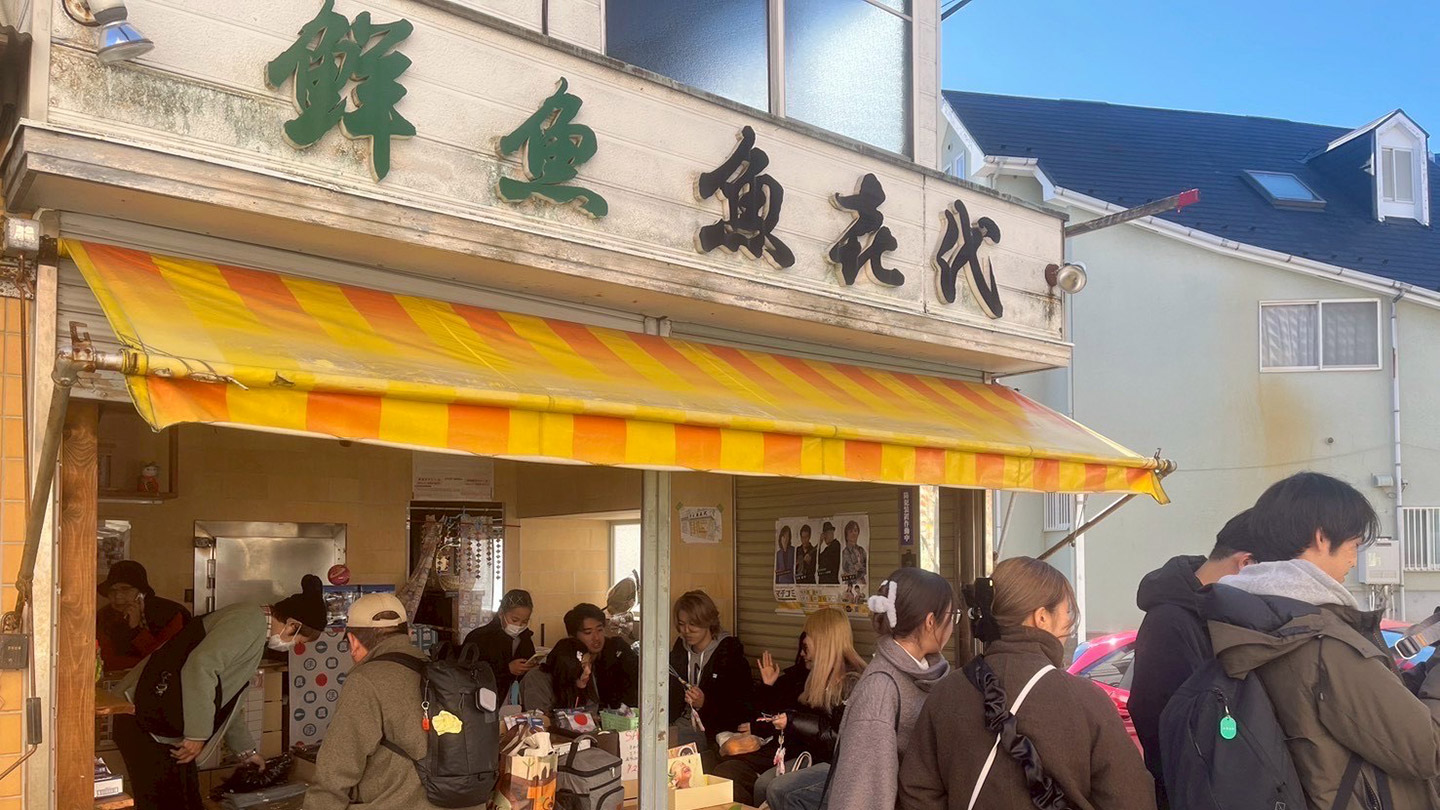 鮮魚 魚㐂代」と書かれた看板を掲げた建物。なかにはベンチが置かれ、多くの人が集まっている