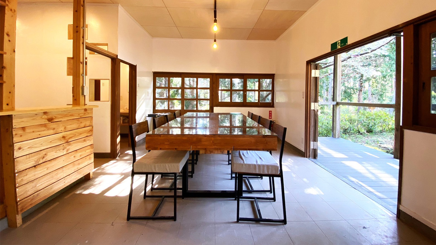 窓から外光が差し込む明るいダイニングルーム。部屋の中央には長い木製テーブルと椅子が並び、大きく開放された扉の外には緑の木々が見える