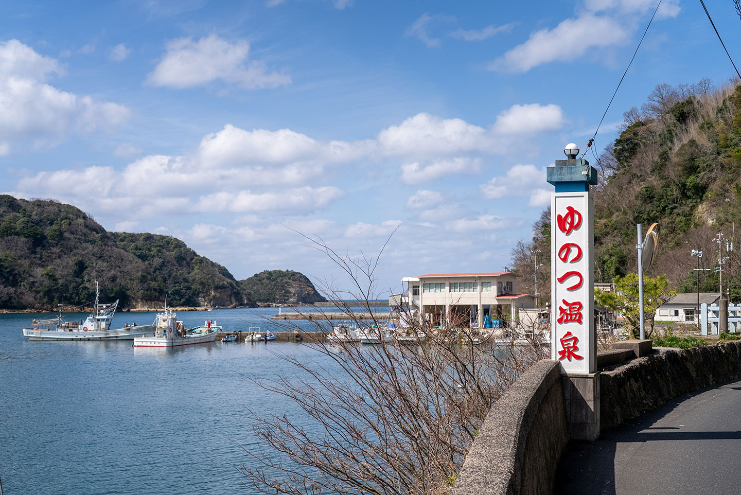 青空の下に広がる島根県大田市温泉津の港の風景。画面右側には「ゆのつ温泉」と赤い文字で書かれた白い看板が立ち、左側の穏やかな海には白い漁船が数隻停泊している