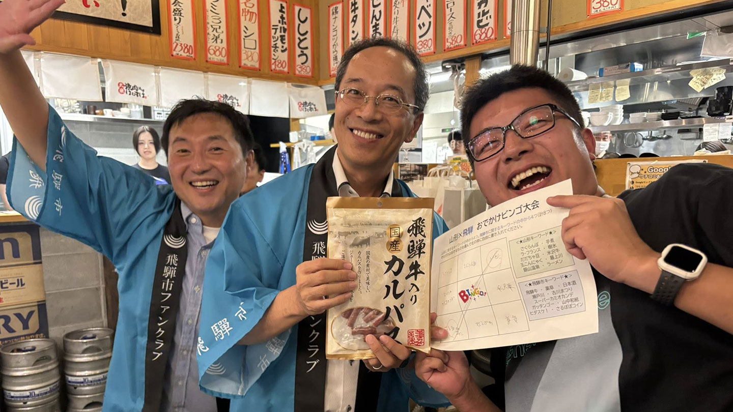 法被を着た男性2名と、黒いジャケットを着て紙を持つ男性が並ぶ