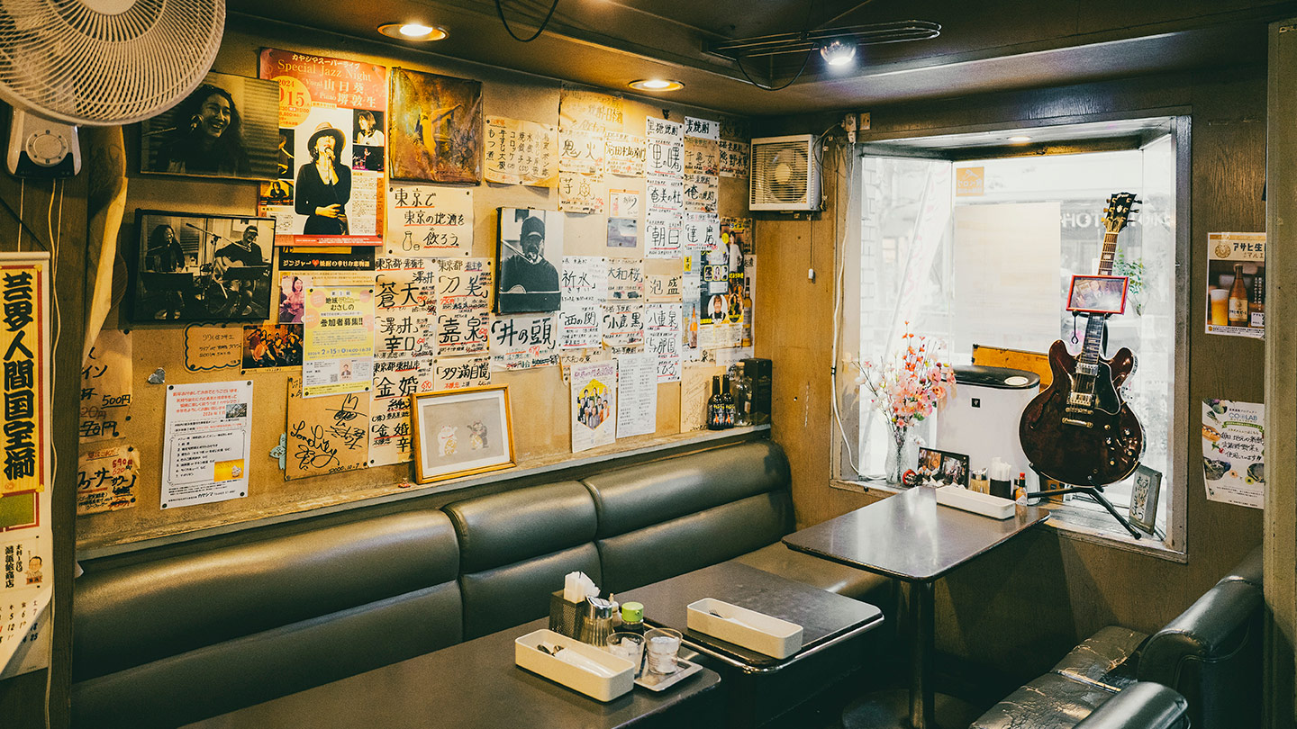 壁一面にポスターや手書きメニューが貼られた喫茶店の店内。ソファ席とテーブルが並び、窓辺にはギターが飾られている