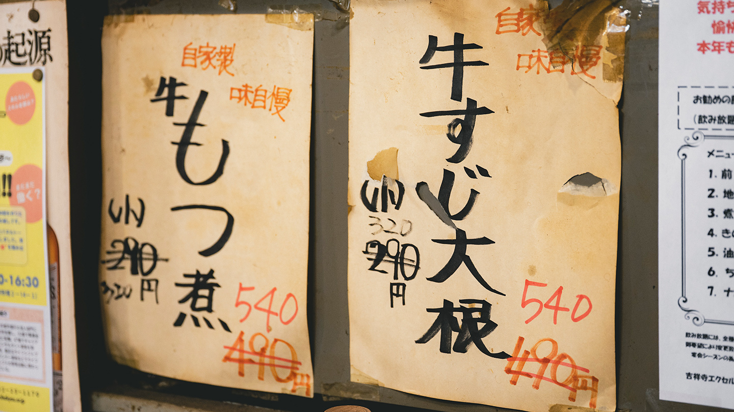 店内の壁に貼られた手書きの料理メニュー。「牛もつ煮」や「牛すじ大根」などの価格が書かれた紙が並ぶ
