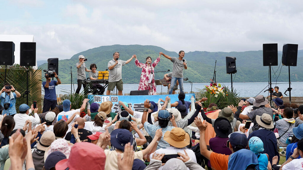 船浮音祭りの様子。舞台にいる三人が手をつなぎながら上げている。観客も手をあげて盛り上がる