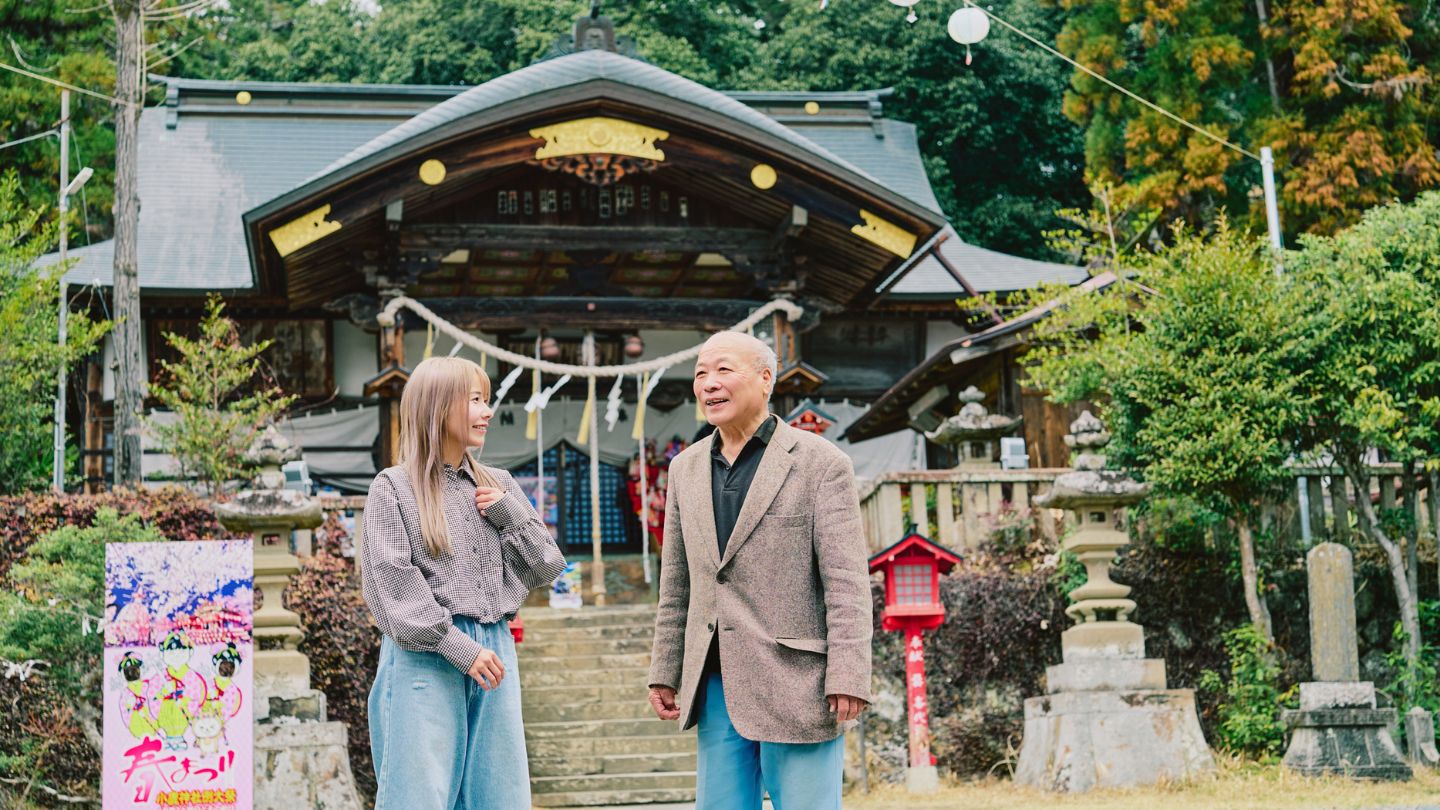 神社の本殿を背景に話をするこつぶさんと宮司さん