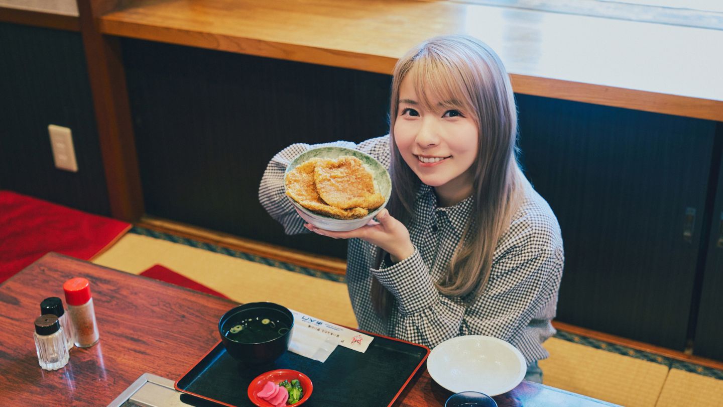 店内の座敷にて、顔のサイズほどある大きな「わらじカツ丼」を掲げて笑顔を見せるこつぶさん