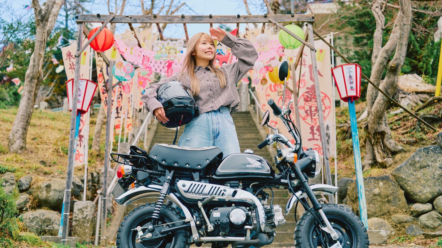 ひな祭りの装飾がある神社の階段を背景に、バイクの後ろでポーズをとるこつぶちゃんねるさん
