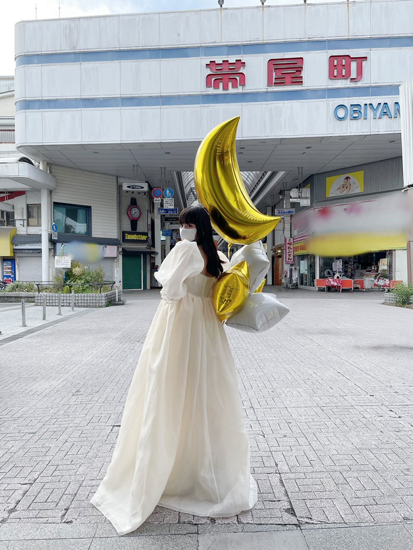 帯屋町商店街のアーケード前で、白い衣装を着た鶴嶋乃愛さんが金色の月型バルーンを背負って立っている