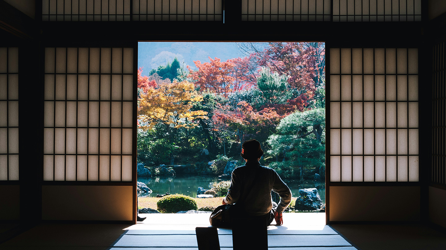 和室の開口部から、池と紅葉した木々が広がる日本庭園を眺める人物の後ろ姿